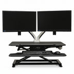 An electric standing desk converter with a black tabletop, supporting two monitors, a keyboard, and a mouse. It has an integrated USB charging port and is designed for minimal assembly.